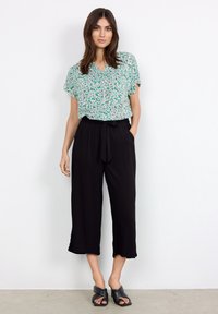 Blusa corta de manga corta verde y blanca con estampado floral, combinada con pantalones de sastre negros de cintura alta y pierna ancha con un cinturón atado; sandalias negras tipo slide.