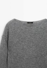 Pull gris en tricot avec un col rond et une étiquette visible au niveau du col sur un fond blanc uni.