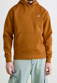 Hombre vestido con una sudadera marrón con el logo Jumpman blanco y unos pantalones cargo azul claro, de pie frente a un fondo liso.