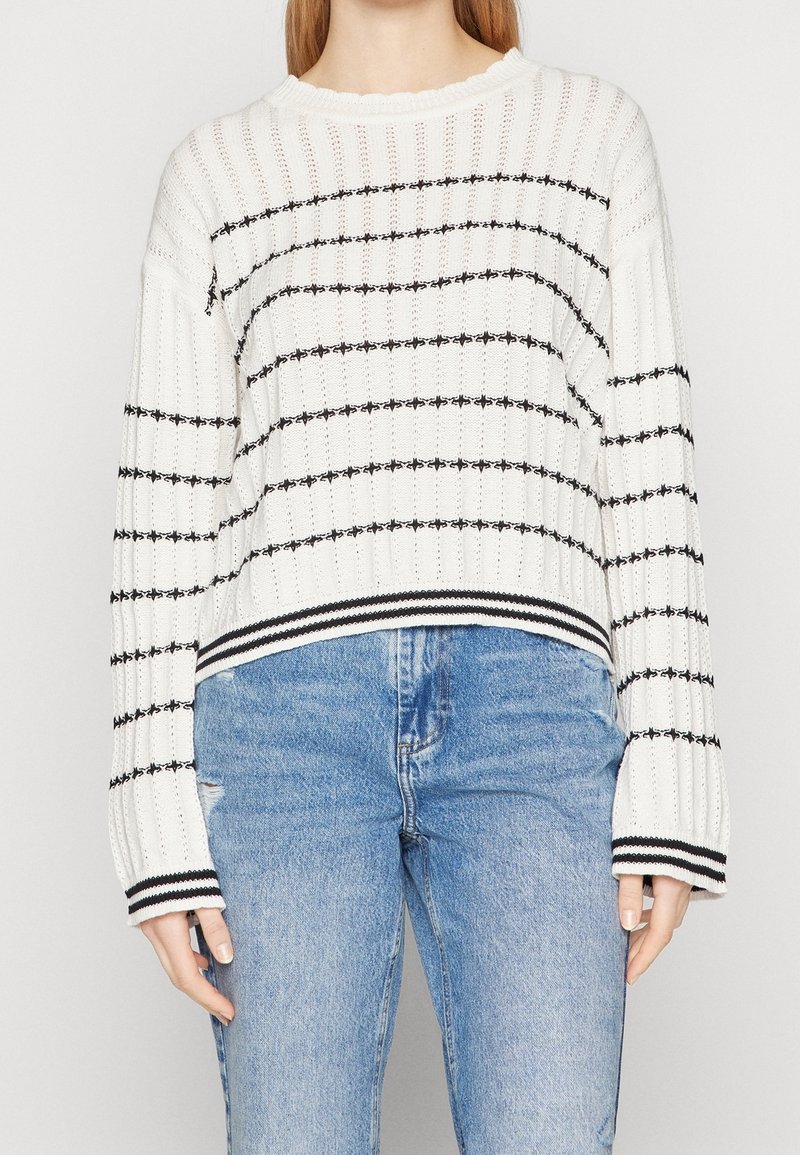 Biały dziany sweter z czarnymi poziomymi paskami, prążkowaną fakturą i szerokimi rękawami. Posiada paskowany dół i luźny fason.