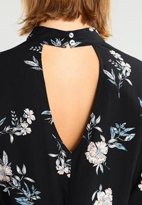 Schwarzes Blumenkleid mit V-förmigem Rückenausschnitt, kleiner Knopfleiste am Ausschnitt und hellrosa-blauen Blumenmustern.