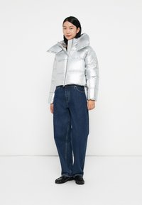 Silver puffer jacket with a high collar, zip front, and horizontal quilted texture, paired with loose blue denim jeans and black shoes.