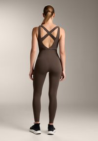 Brown fitted jumpsuit with cross-back straps and a low cut back. Smooth texture, full-length legs, paired with black athletic shoes.