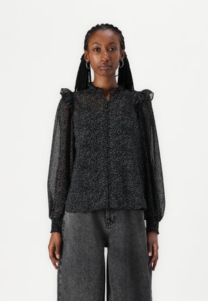Blusa nera con pois bianchi sparsi, maniche lunghe trasparenti, spalle arricciate e chiusura con bottoni davanti. Tessuto testurizzato con una leggera lucentezza.