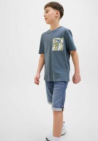 T-shirt gris à manches courtes avec une poche à motifs, shorts en denim bleu retroussés aux poignets et baskets blanches. Coupe décontractée, matériau léger.