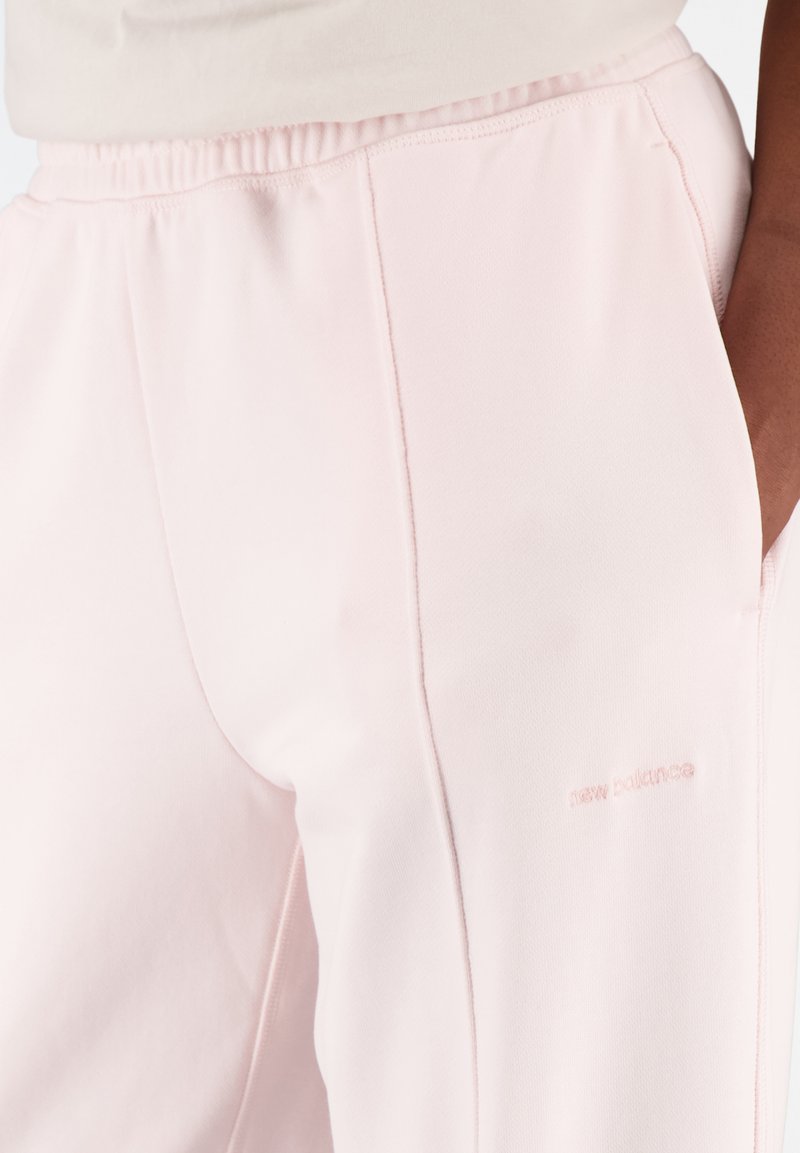 Pantalons de survêtement New Balance rose clair avec poches latérales portés par une personne, montrant une ceinture élastique et un logo discret sur la cuisse.