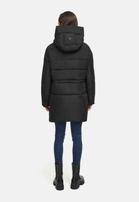 Black puffer jacket with a hood, featuring horizontal quilted lines, a slightly cinched waist, and a logo on the back. Worn with blue jeans and black boots.