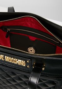 Black quilted tote bag with gold hardware. Interior features a red lining, a zip pocket, and a branded card holder.