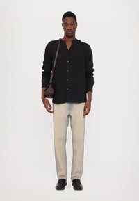 Camicia nera con bottoni, pantaloni beige a gamba dritta, scarpe nere e borsa marrone piccola. La camicia ha una vestibilità comoda e un design senza colletto.