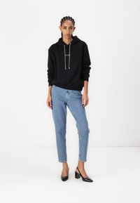 Svart hettegenser med snorer og tekst foran, kombinert med høyhårede lyseblå jeans og svarte lakkhæl. Enkel design.