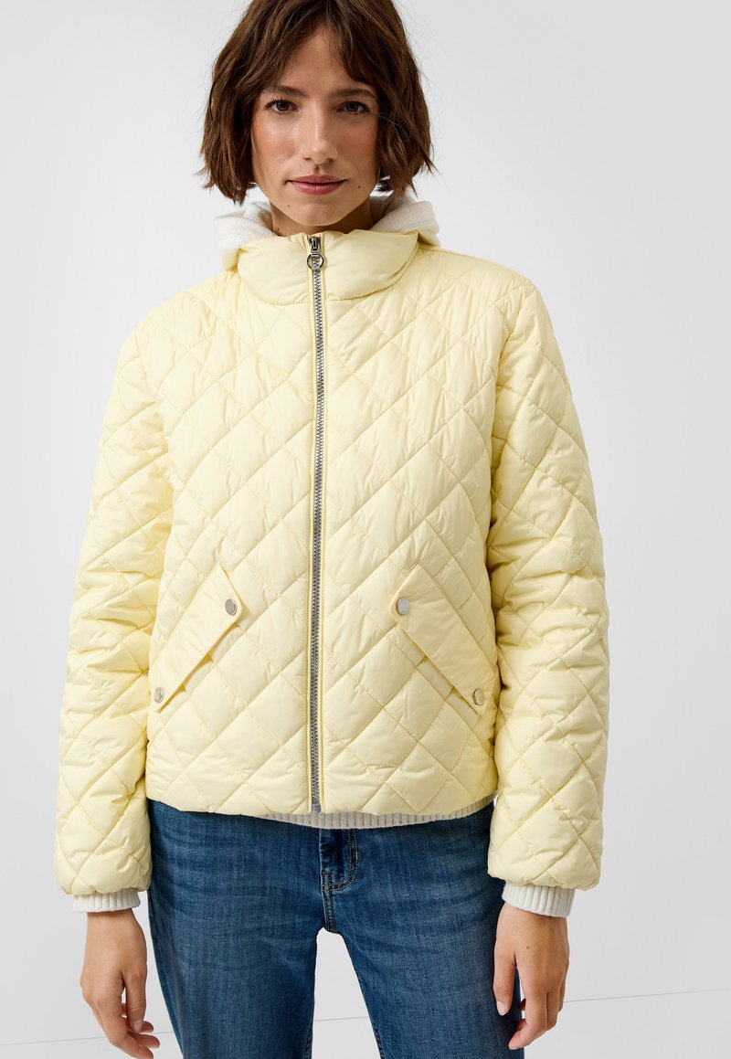 Frau trägt eine hellgelbe Steppjacke mit diagonaler Steppung und vorderen, mit Knöpfen versehenen Klappentaschen, kombiniert mit blauen Jeans.