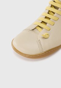 Scarpa in pelle beige con finitura testurizzata, lacci in tessuto giallo e punta arrotondata. La suola presenta cuciture e un bordo leggermente elevato.