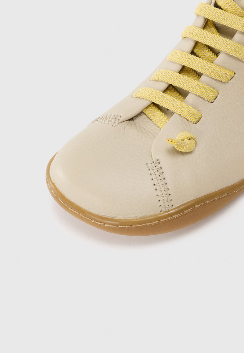 Chaussure en cuir beige avec une finition texturée, lacets en tissu jaune et bout arrondi. La semelle présente des coutures et un bord légèrement surélevé.