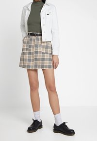 Chaqueta de mezclilla blanca, suéter de cuello alto oliva, falda de cuadros beige con bolsillos, zapatos negros de cordones y calcetines blancos a rayas.
