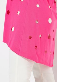 Camicia fucsia con grandi ritagli rotondi, lati corti che si assottigliano verso il basso, abbinata a pantaloni bianchi, dal tessuto liscio e dal taglio casual.