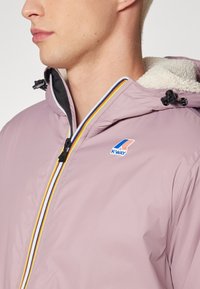 Profil latéral d'une personne portant une veste K-Way violet clair avec une fermeture éclair rayée multicolore et une capuche doublée en polaire.