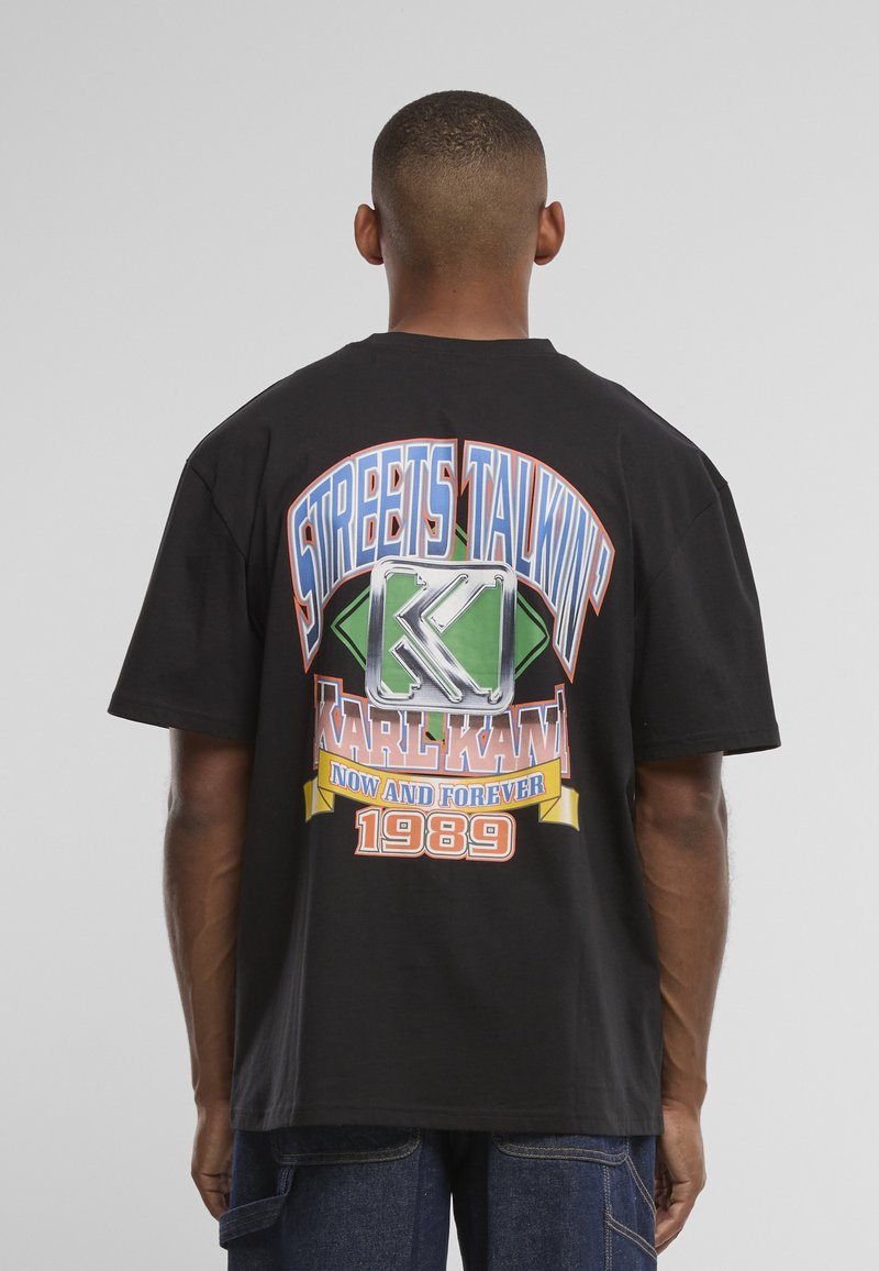Άνδρας που φοράει μαύρο oversized T-shirt με πολύχρωμη εκτύπωση "Streets Talkin'," "Karl Kani," "Now and Forever" και "1989" στην πλάτη.