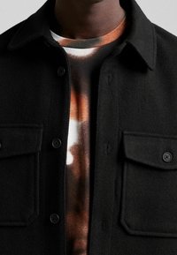 Detailní záběr na osobu v černém zapínacím kabátu přes hnědé a bílé tričko s batikovaným vzorem, zaměřený na horní část trupu a výstřih.