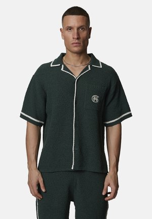 Homme portant une chemise à manches courtes vert foncé avec des bordures blanches et un pantalon assorti, debout devant un fond uni.