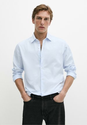 Giovane uomo con capelli castano chiaro indossa una camicia azzurra con bottoni e maniche arrotolate e jeans neri, in piedi con le mani in tasca.