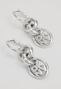 Silver hoop earrings featuring intertwined links and a circular charm with the engraved text "DKNY." Smooth, shiny surface with a modern design.