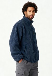 Veste polaire bleue avec col montant, fermeture éclair intégrale et deux poches latérales. Présente un tissu texturé et un panneau lisse et contrastant sur une épaule.