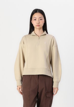 Beige zip-up hette laget av mykt stoff, med en liten logo, ribbestrikkede mansjetter og en cropped design. Kombinert med mørkebrune cargo-bukser.