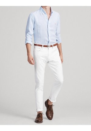 Mann trägt hellblau gestreiftes Hemd mit Knöpfen, weiße Slim-Fit-Jeans mit braunem Gürtel und braune Leder-Herrenschuhe, steht vor einem schlichten Hintergrund.