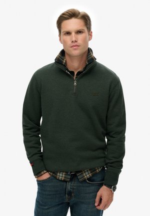 Grüner Fleece-Pullover mit einem Reißverschlusskragen, über einem schwarz-gelben Karobluse. Blaue Jeans mit einer Uhr, die am Handgelenk sichtbar ist.