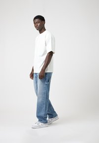 T-shirt blanche à manches courtes associée à un jean bleu clair. Les chaussures sont grises et blanches. L'arrière-plan est simple et clair.