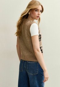 Maglione senza maniche marrone in maglia con finitura testurizzata e motivo a quadri neri, indossato sopra una camicia bianca, abbinato a jeans blu a vita alta.