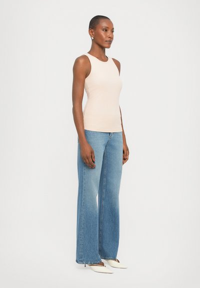 Beige ribbed tank top with a crew neckline paired with light blue flared jeans. Model stands in a neutral background. White pointed-toe heels.