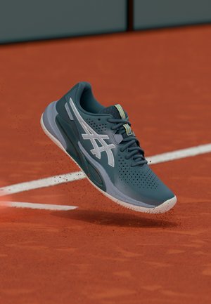 Blue and white ASICS tennis shoe mid-air above red clay court, kicking up small dirt particles near white boundary lines.