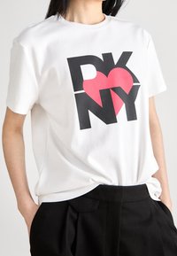 Biała bawełniana koszulka z krótkim rękawem z wyrazistym czarnym napisem "DKNY" i czerwonym sercem, o luźnym kroju. Noszona z czarnymi spodniami.