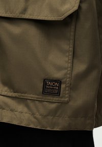 Κοντινή λήψη ελιάς πράσινου υφάσματος με ραμμένη ρέλια και μαύρη ετικέτα με την ένδειξη «TAION» και κείμενο αποστολής προϊόντος.