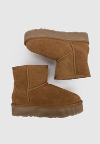 Botas de tobillo de ante marrón con suelas de goma gruesa y estriada, costuras visibles y puntas redondeadas suaves sobre un fondo claro y liso.