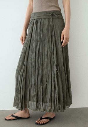 Kvinde iført ankel-lang, krøllet olivengrøn nederdel med taljebånd og sorte flip-flops, stående mod en ensfarvet baggrund.