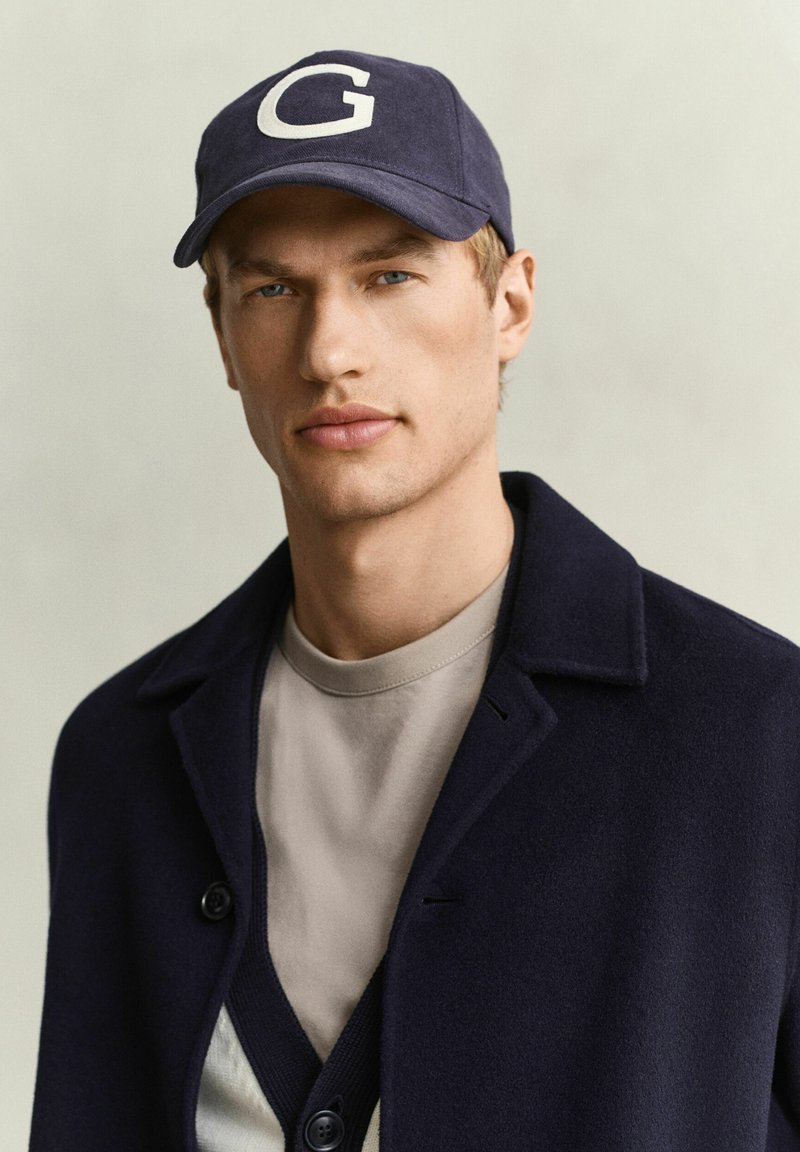 Jeune homme portant une casquette bleu marine avec un "G," blanc, un manteau foncé et une chemise gris clair, regardant directement la caméra.