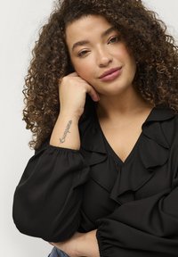 Schwarze Bluse mit Rüschendetails, gerafften Ärmeln und V-Ausschnitt. Lockige Haare und ein Unterarmtattoo im Vordergrund sichtbar.