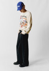 Una persona che indossa un cappellino blu KTM, un maglione color crema con grafiche colorate, pantaloni neri larghi con cuciture bianche e scarpe nere, in piedi con le mani nelle tasche.