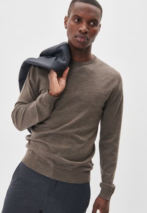 Maglione marrone a maglia con maniche lunghe, colletto e orlo a costine. Indossato con un blazer blu scuro appoggiato sulla spalla. Sfondo semplice.