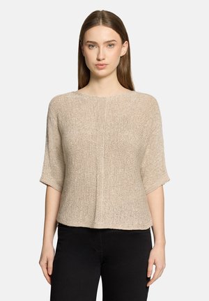 Vrouw met een beige, kort mouw gebreide trui en zwarte broek, staande tegen een effen lichte achtergrond, met het gezicht naar voren gericht.