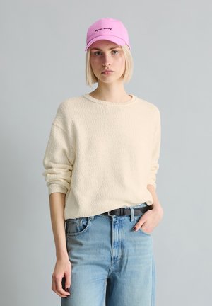 Jeune personne portant une casquette rose avec texte, un pull crème texturé, un jean bleu clair, debout avec une main dans la poche sur un fond uni.