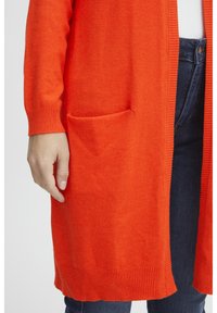 Long cardigan orange en tissu doux, avec des poignets et un ourlet côtelés, et une poche unique sur le bas à l'avant.