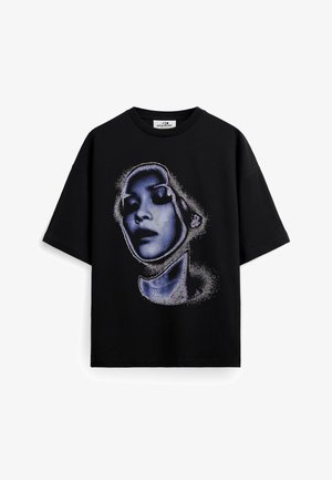 Młody mężczyzna z krótkimi włosami stoi, ubrany w czarny, oversize'owy t-shirt z niebieskim artystycznym wzorem twarzy oraz białe luźne spodnie, ręce w kieszeniach.