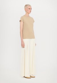 Beige tričko s krátkým rukávem a jemným potiskem, v kombinaci s volnými, krémovými širokými kalhotami, stojící na minimalistickém pozadí.