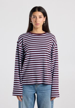 Vrouw met lang donker haar draagt grote oorringen, een bordeauxrood en lichtblauw horizontaal gestreept shirt met lange mouwen en blauwe spijkerbroek tegen een effen achtergrond.