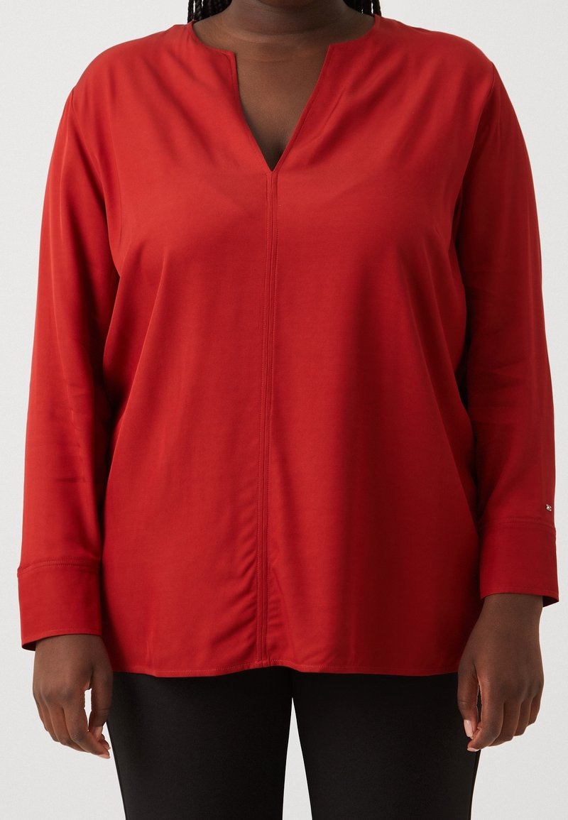 Blouse rouge à manches longues avec un décolleté en V, fabriquée dans un tissu lisse, avec des coutures latérales et des manches à poignets. Design simple sans motifs.