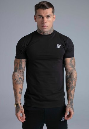 Homme musclé avec des tatouages au cou et aux bras, portant un T-shirt noir ajusté et un pantalon noir, debout devant un fond gris uni.