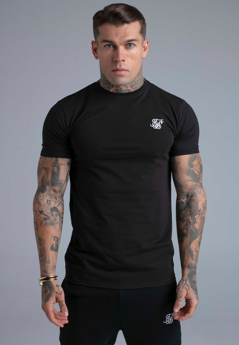 Homme musclé avec des tatouages au cou et aux bras, portant un T-shirt noir ajusté et un pantalon noir, debout devant un fond gris uni.